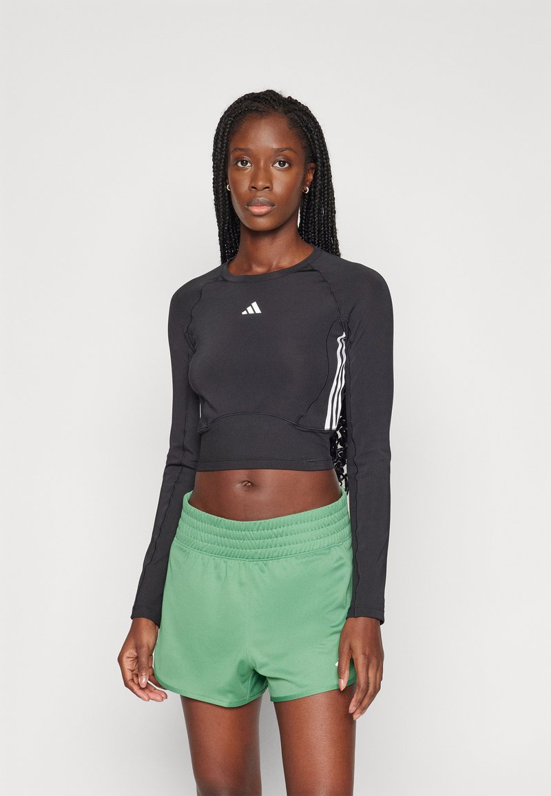 Schwarzes, langärmliges, verkürztes Oberteil mit geripptem Design und weißem Adidas-Logo, kombiniert mit grünen Sportshorts mit elastischem Bund.