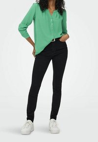 Donna con una blusa verde abbottonata, pantaloni neri attillati, sneakers bianche, in piedi con una mano in tasca su sfondo bianco.