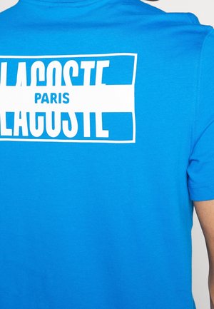 Lyseblå skjorte med hvidt rektangulært logo, der viser ordene "Lacoste" og "Paris" centreret inde i logoet på den øvre ryg.