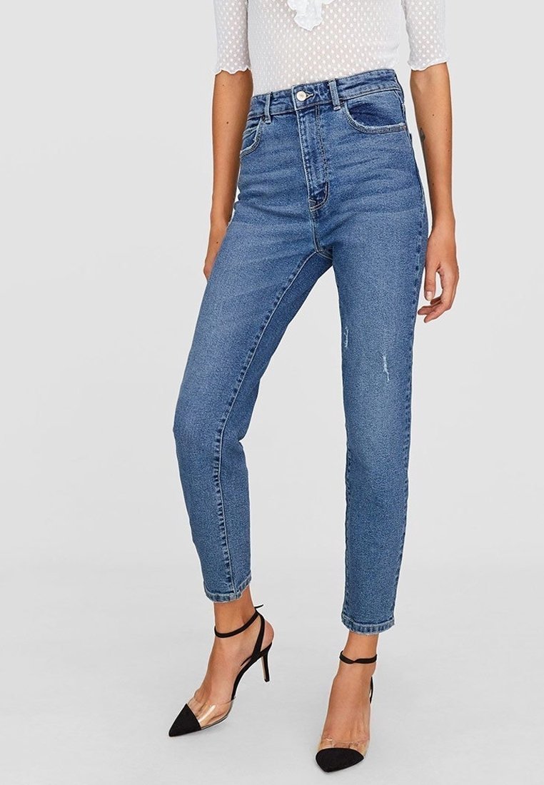 Frau in hoch taillierten, blauen Skinny-Jeans mit leichten Abschürfungen, einem transparenten weißen Oberteil und schwarzen hochhackigen Schuhen mit Knöchelriemen.