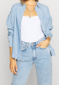 Kobieta w jasnoniebieskich dżinsach, białej bluzce na ramiączkach, rozpiętej jasnoniebieskiej koszuli dżinsowej i warstwowych złotych naszyjnikach, z jedną ręką w kieszeni.