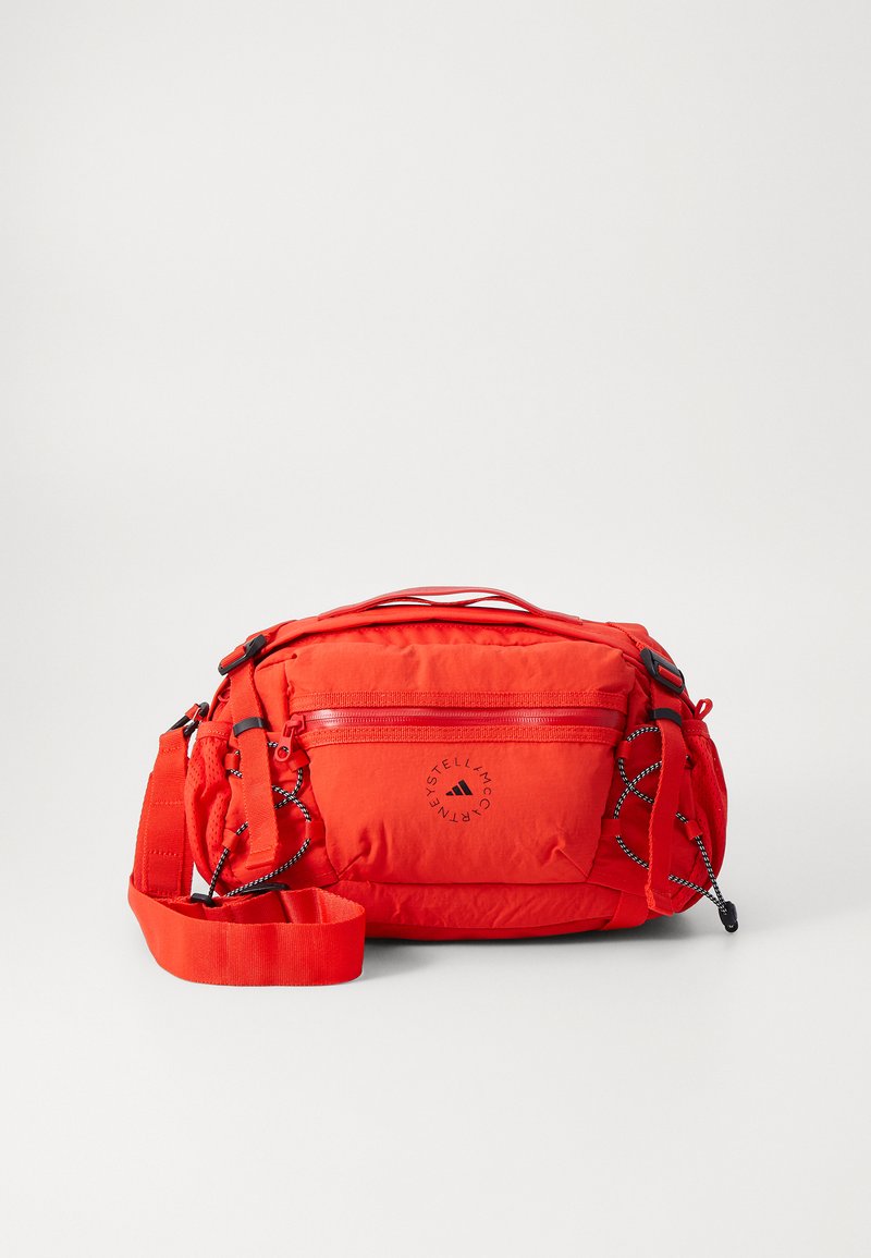 Červená nylonová crossbody taška s nastavitelným popruhem, přední zipová kapsa a logo detail. Obsahuje texturované akcenty a kontrastní černé kovové části.