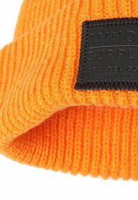 Beanie arancione lavorato a maglia con una texture a coste. Presenta una toppa in pelle nera cucita sul tessuto.