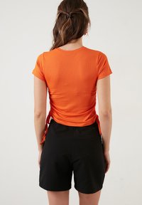 Oranje geribbelde top met korte mouwen en een strikdetail aan de zijkant, gecombineerd met zwarte shorts, gezien van achteren.
