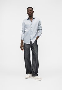 Hellblaues Hemd mit Knopfleiste und feinem Streifenmuster, kombiniert mit locker sitzenden dunkelblauen Jeans und weißen Sneakers mit beigefarbenen Akzenten.