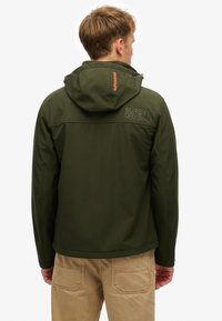 Veste softshell vert foncé avec capuche, ornée d'une broderie de marque dans le dos et d'un détail d'étiquette orange contrastante près du col.