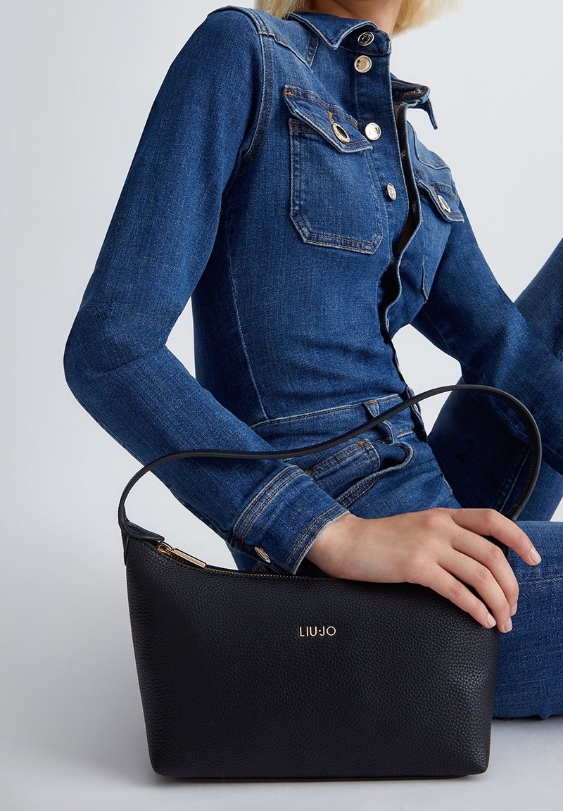 Femme en tenue en denim tenant un sac à main Liu Jo en cuir texturé noir avec fermeture éclair dorée et logo à l'avant, sur fond neutre.