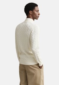 Svetrový pulóver s vrkočovým vzorom v krémovej farbe s vysokým golierom a rebrovanými manžetami. Textúrovaná látka a voľný strih kontrastujú s béžovými nohavicami.