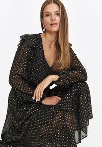 Czarna przezroczysta sukienka w złote kropki, z falbanami przy rękawach i dekoltem w kształcie litery V. Lejący się krój, teksturowany materiał i szczegóły w postaci warstwowej spódnicy.