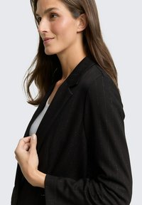 Zwarte blazer met subtiele krijtstrepen, lichte glans, notch-revers en een getailleerde pasvorm. Model toont een ontspannen handpositie.