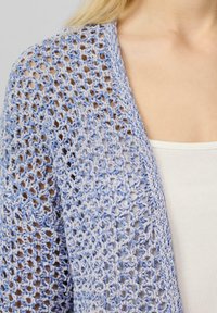 Hellblaues, offen gestricktes Cardigan mit gehäkeltem Design, weicher Textur und drapiertem Schnitt. Über einem weißen Trägershirt getragen.