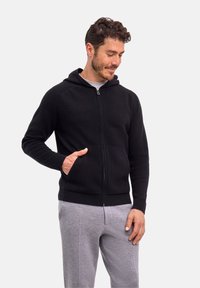 Schwarzer Kapuzenpullover mit Reißverschluss und strukturierter Strickoptik, ausgestattet mit einer Fronttasche und einer Kapuze. Darüber wird ein graues T-Shirt getragen, kombiniert mit hellgrauen Hosen.