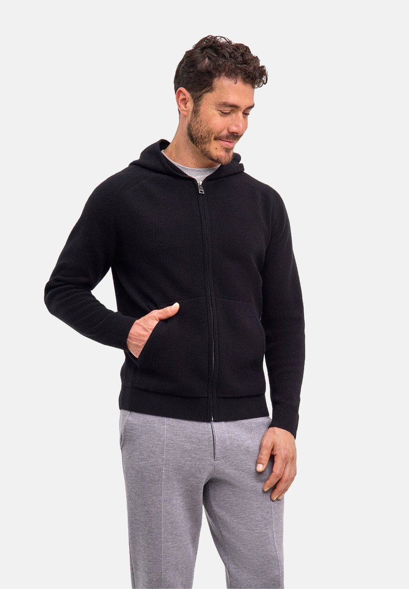 Schwarzer Kapuzenpullover mit Reißverschluss und strukturierter Strickoptik, ausgestattet mit einer Fronttasche und einer Kapuze. Darüber wird ein graues T-Shirt getragen, kombiniert mit hellgrauen Hosen.