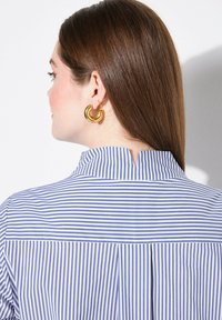 Blusa a righe blu e bianche con colletto alto e dettaglio spaccato sul retro. Il modello indossa orecchini a cerchio dorati, con capelli lisci e lucenti.