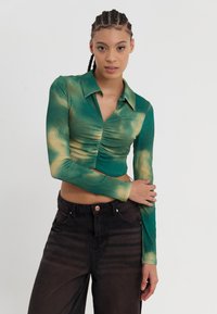 Vihreä ja keltainen tye-dye pitkähihainen lyhyt toppi, jossa on kauluspaita ja rypytysdetaljeja. Yhdistetty tummanmustiin farkkuihin.