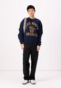 Sweatshirt azul marinho com gola redonda e impressão gráfica dourada com "Santa Monica Grizzlies" e leão. Usado com calças cargo pretas e ténis brancos.