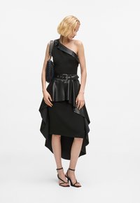 Μαύρο φόρεμα με έναν ώμο και στρωτή ασύμμετρη τελείωμα, δερμάτινη ζώνη και μαύρη τσάντα. Σανδάλια με λουριά ολοκληρώνουν την εμφάνιση.