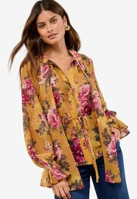 Blumenbluse mit langen Ärmeln, die einen senfgelben Hintergrund, rosa Rosen und eine Bindewasit hat. Ausgestellte Manschetten verleihen Textur und Detail.