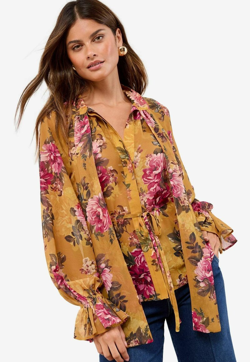 Blumenbluse mit langen Ärmeln, die einen senfgelben Hintergrund, rosa Rosen und eine Bindewasit hat. Ausgestellte Manschetten verleihen Textur und Detail.