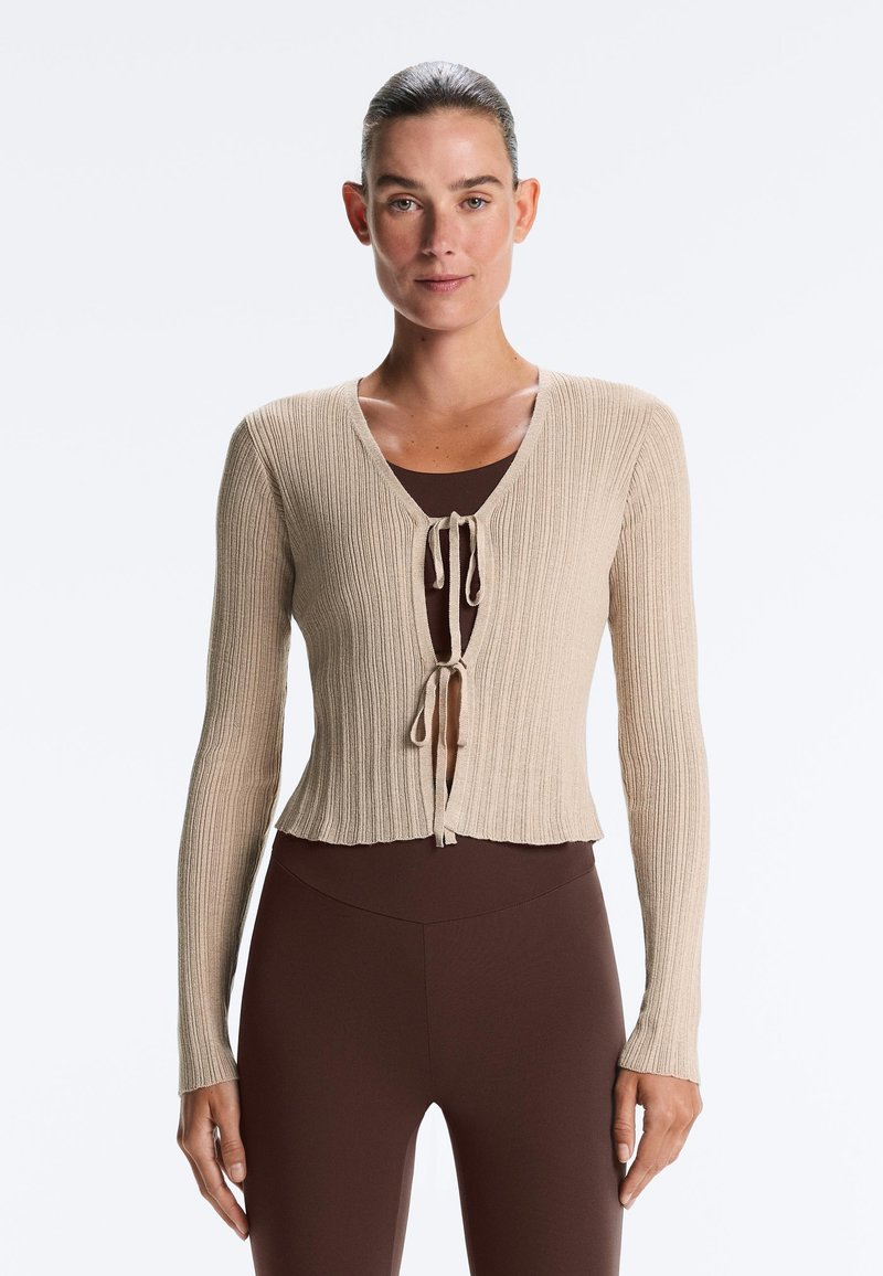 Femme aux cheveux coiffés en arrière portant un cardigan côtelé beige noué deux fois sur un haut marron et un pantalon marron ajusté, debout devant un fond blanc.