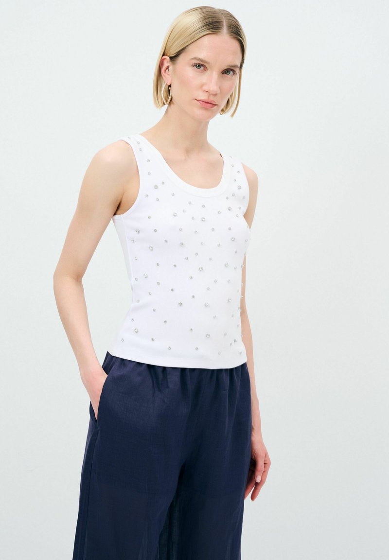 Top bianco senza maniche con scollatura rotonda, decorato con piccole embellishments argentate, abbinato a pantaloni larghi blu navy. Tessuto dalla texture liscia.