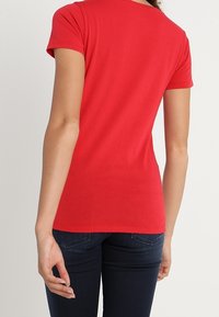 Mujer con una camiseta ajustada de manga corta roja y jeans oscuros, vista desde la espalda contra un fondo claro.