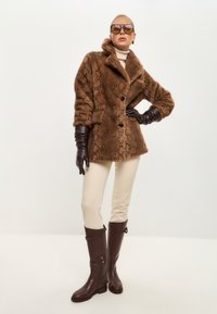 Brauner Kunstpelzmantel mit Schlangenmustern, cremfarbener Rollkragenpullover, beige enganliegende Hose, braune Lederhandschuhe und kniebundhohe braune Stiefel mit goldenen Reißverschlüssen.