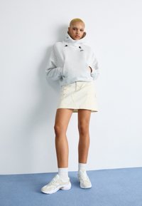 Šedá mikina s kapucí, krémová sukně s předními kapsami, bílé tenisky s jemnými akcenty. Pozadí je světle modré a bílé.