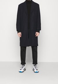 Marineblauer Wollmantel mit einem langen, lässigen Schnitt, versehen mit einem einzigen Knopfverschluss, kombiniert mit schwarzen Hosen und weißen Sneakers mit blauen Akzenten.