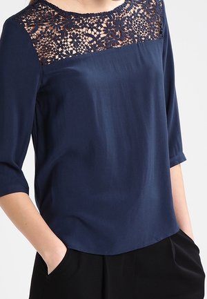 Marinebluse med blonder over skuldrene, 3/4-ærmer og en glat tekstur, kombineret med sorte bukser.