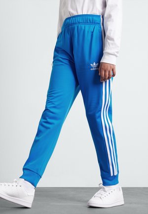 Person iført lyseblå Adidas træningsbukser med hvide striber og hvide sneakers, stående på gråt gulv mod ensfarvet baggrund.