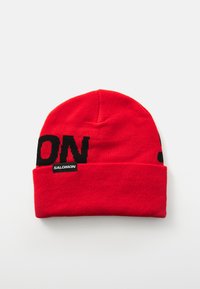 Rode gebreide beanie met een zwart blokletterontwerp met "SALOMON." Het materiaal lijkt zacht; het ontwerp heeft een omgevouwen rand.