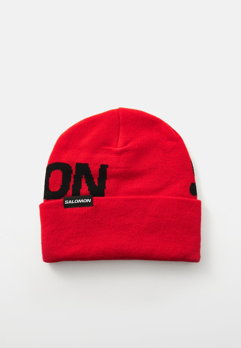 Rode gebreide beanie met een zwart blokletterontwerp met "SALOMON." Het materiaal lijkt zacht; het ontwerp heeft een omgevouwen rand.