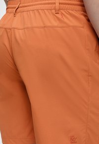 Κοντινή λήψη πορτοκαλί σορτς με ελαστική μέση και ετικέτα ηλιακής προστασίας UPF 50+ SolarPack στο κάτω μέρος του ποδιού.