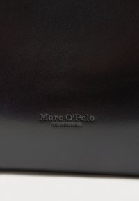 Crni kožni materijal glatke teksture s utisnutim imenom marke "Marc O'Polo" i detaljem osnivanja "EST. U STOCKHOLMU."