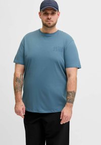 Blaues Baumwoll-T-Shirt mit kurzen Ärmeln, rundem Ausschnitt und einem dezenten, geprägten Logo auf der Brust. Kombiniert mit schwarzen Hosen.