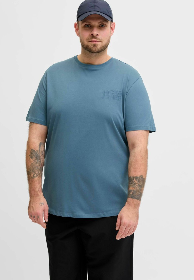 Blaues Baumwoll-T-Shirt mit kurzen Ärmeln, rundem Ausschnitt und einem dezenten, geprägten Logo auf der Brust. Kombiniert mit schwarzen Hosen.