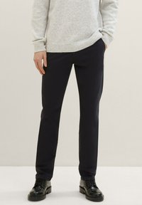 Pantaloni stretch blu navy a vestibilità slim; dotati di tasche laterali, una texture liscia, abbinati a un maglione in maglia grigio chiaro e scarpe nere.