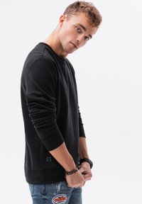Schwarzer Pullover mit langen Ärmeln und gerippten Bündchen, ausgestattet mit einem Rundhalsausschnitt und strukturiertem Stoff. Getragen mit abgetragenen Jeans.