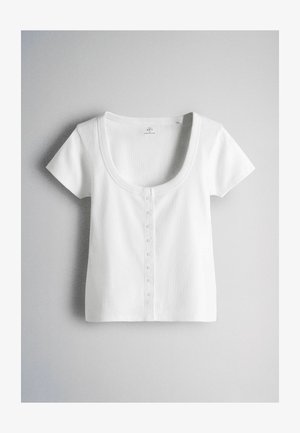 Rövid ujjú, fehér bordázott crop top mély kivágással és apró első gombokkal világos háttéren.