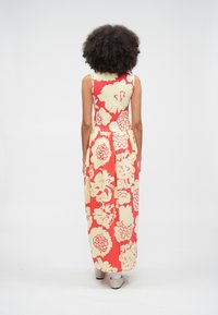 Žena s kučeravými vlasmi v červených a krémových kvetovaných maxi šatách bez rukávov stojaca chrbtom na jednobielom pozadí.