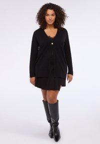 Schwarze Strickjacke mit V-Ausschnitt und Knopfverschluss vorne. Weiches Material mit langen Ärmeln, kombiniert mit einem schwarzen Faltenrock und kniehohen Stiefeln.