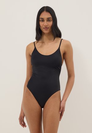 Donna con lunghi capelli scuri che indossa un costume intero nero con spalline sottili, in piedi davanti a uno sfondo chiaro e uniforme.