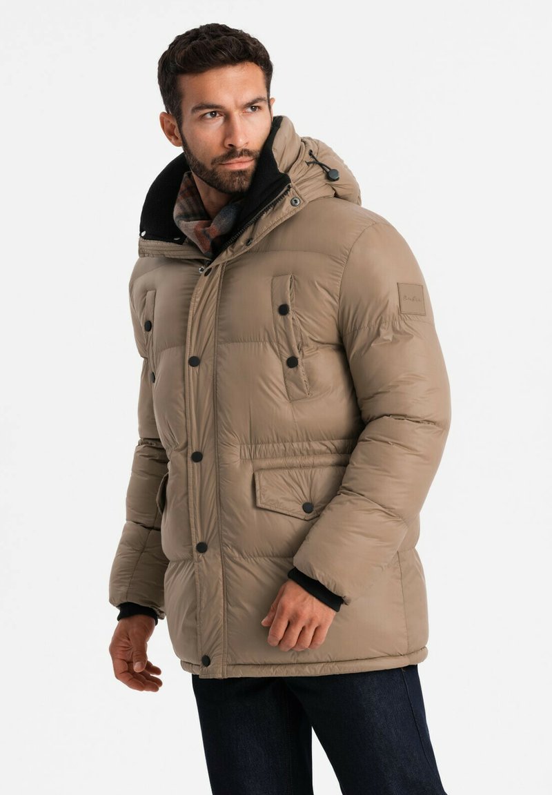 Chaqueta acolchada beige con capucha, presenta botones negros, dos bolsillos frontales y un suave cuello negro. Textura lisa y diseño aislante.