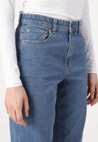 Striped blue denim jeans featuring a high waist, side pockets, and copper-toned hardware. Fabric texture is smooth with vertical white stripes.