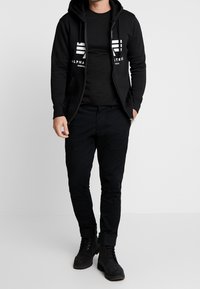 Czarna bluza z kapturem na zamek z białym logo, w połączeniu z czarną koszulką z krótkim rękawem oraz czarnymi spodniami typu slim-fit. Czarną parę butów kończy ten strój.