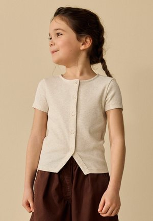 Niña joven con el cabello trenzado, vestida con una camisa beige de manga corta abotonada y pantalones marrones, mirando hacia su izquierda con una leve sonrisa.