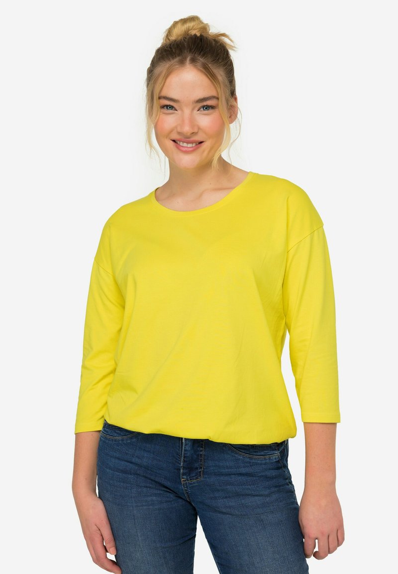 Gelbes Langarmoberteil mit rundem Ausschnitt und lockerer Passform, kombiniert mit dunkelblauen Jeans. Glatte Textur, lässiges Design.