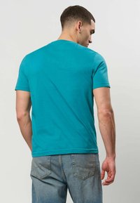 Homme de dos portant un t-shirt turquoise à manches courtes et un jean bleu sur un fond uni clair.