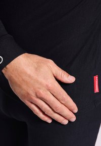 Schwarzes Langarmshirt mit taillierter Passform. Strukturiert sichtbare Stoffoberfläche. Auffälliges rotes Markenlabel an der Seite. Hand ruht auf der Oberfläche des Shirts.